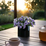 Vase with purple flowers on a wooden table with a cup of coffee and teapot in the foreground, sunset in the background.