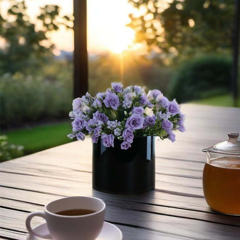 Vase with purple flowers on a wooden table with a cup of coffee and teapot in the foreground, sunset in the background.