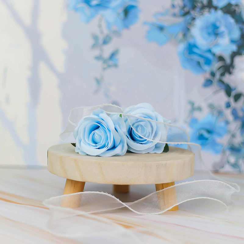 Two blue artificial roses on a small wooden stand with a floral background.