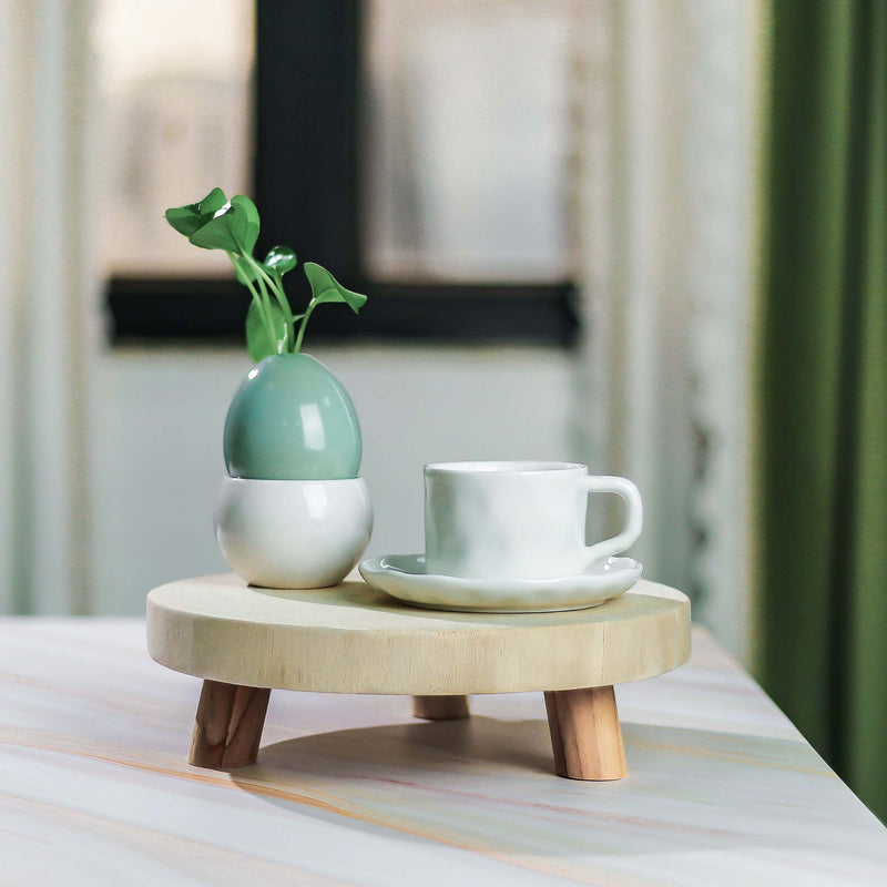 Small wooden table with a white cup, saucer, and green vase on a blurred indoor background