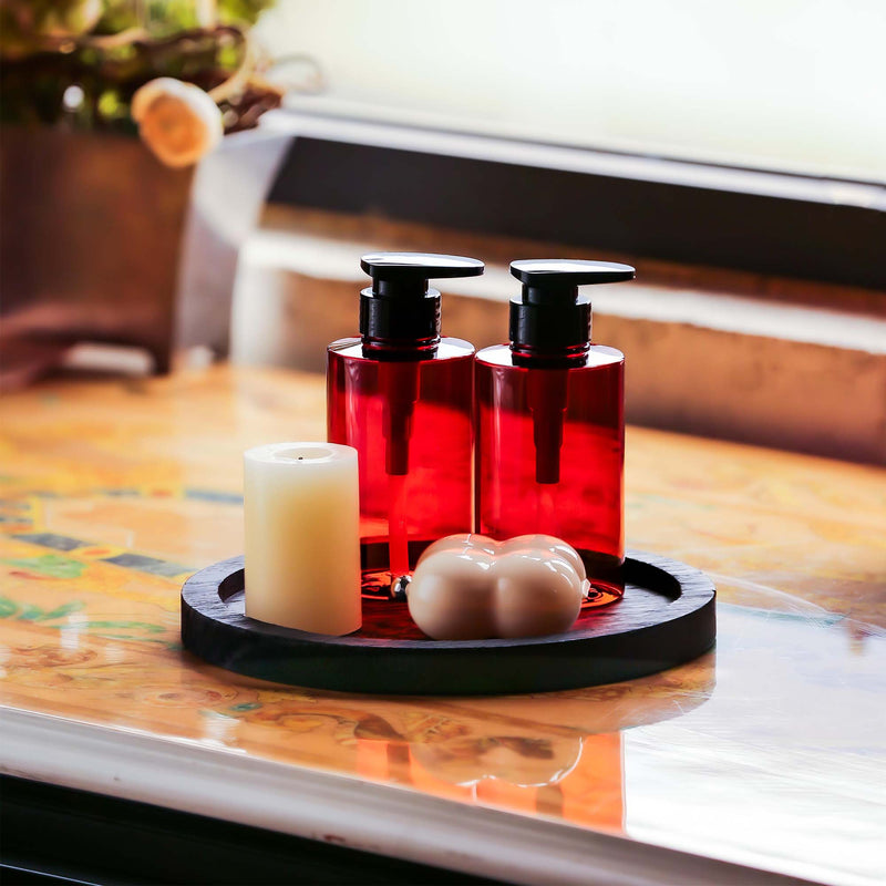 Two red bottles with black pumps on a round black tray, accompanied by a white candle and soap on a wooden surface.