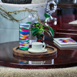 Colorful candle and cup on a wooden coaster with a plant and decorative items in the background.