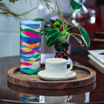 Colorful glass vase on a wooden tray with a white cup and saucer, and a plant in the background.